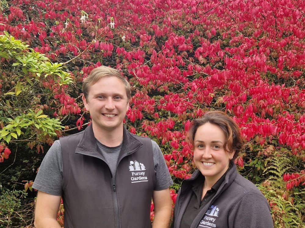 Alex and Abbie Gardening team Young man and woman who are part of the gardening team stood in front of a scarlet leaved bush.