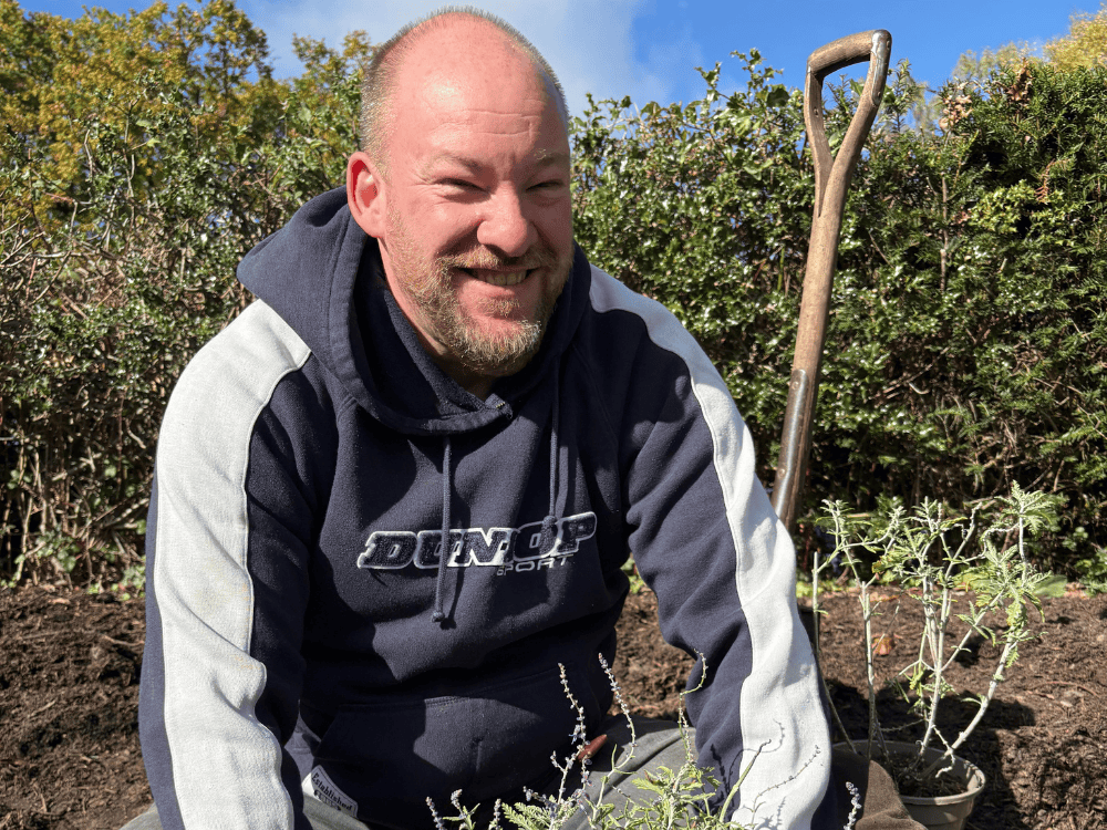 Young man with learning disabilities who is an assistant gardener at Furzey Gardens. Stood in front of a red leaved bush